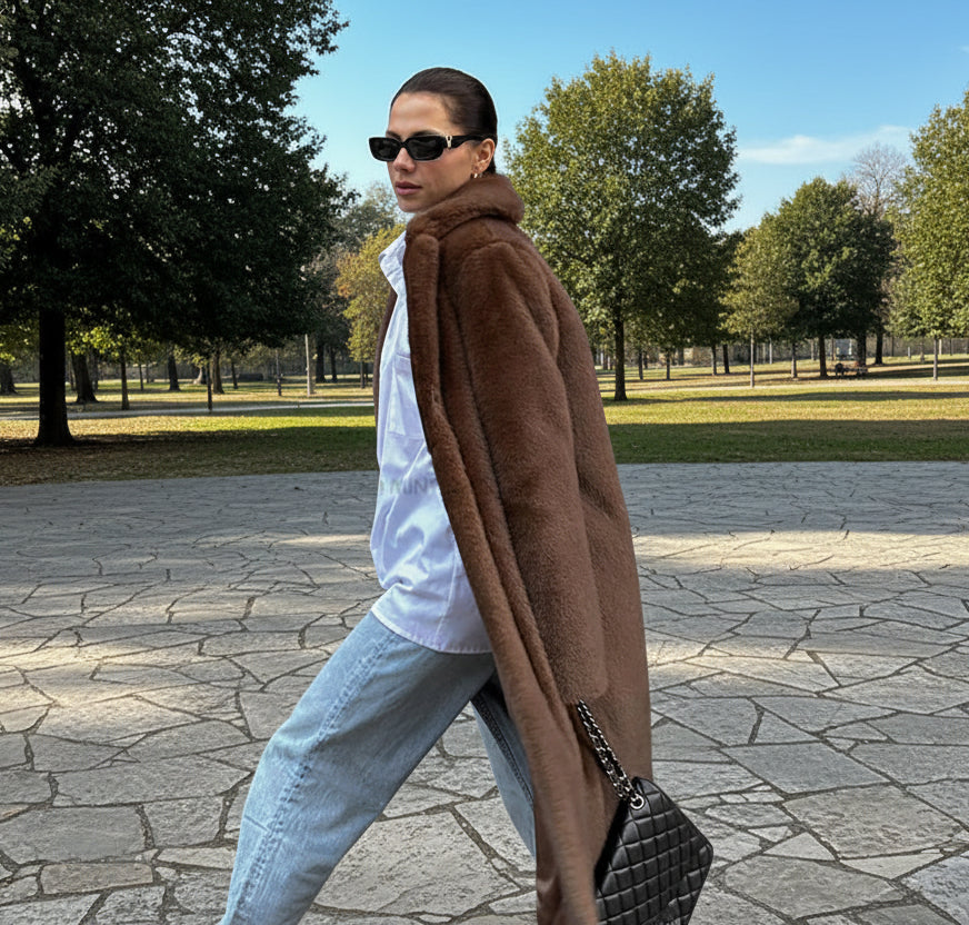 Woman in a brown coat and sunglasses walking past an open park