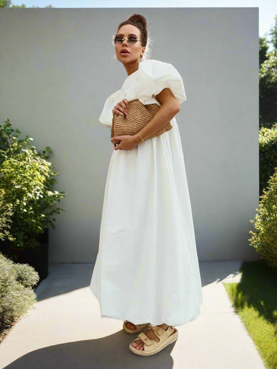Woman in a white dress holding a woven bag against a backyard garden, sunny