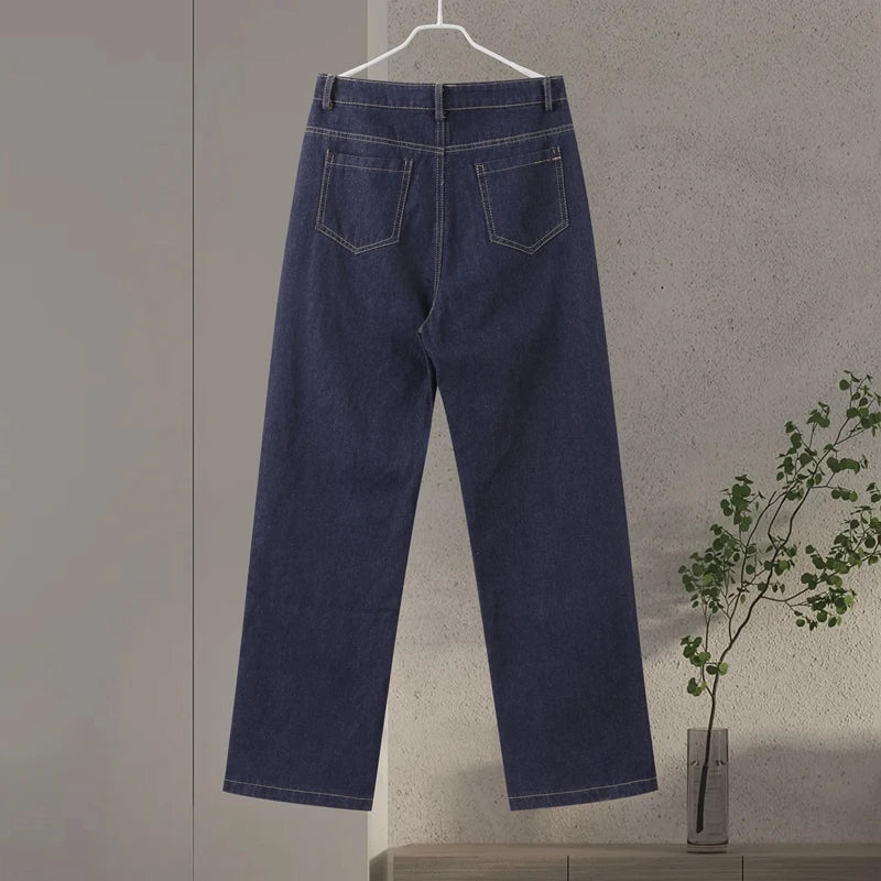 Blue jeans hanging on a hanger against a plain wall with a plant in the corner.