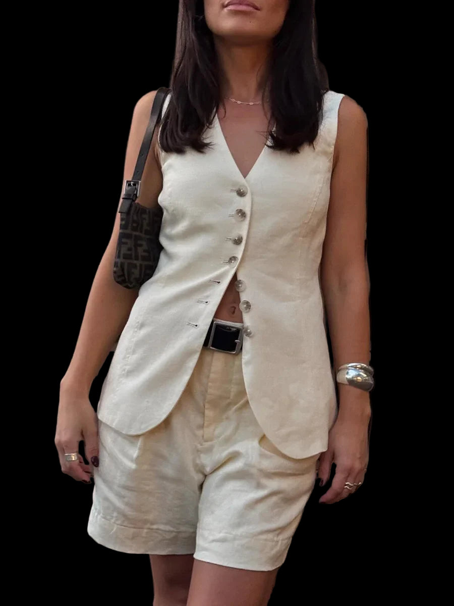Woman wearing a white sleeveless top and beige shorts against a black wall.