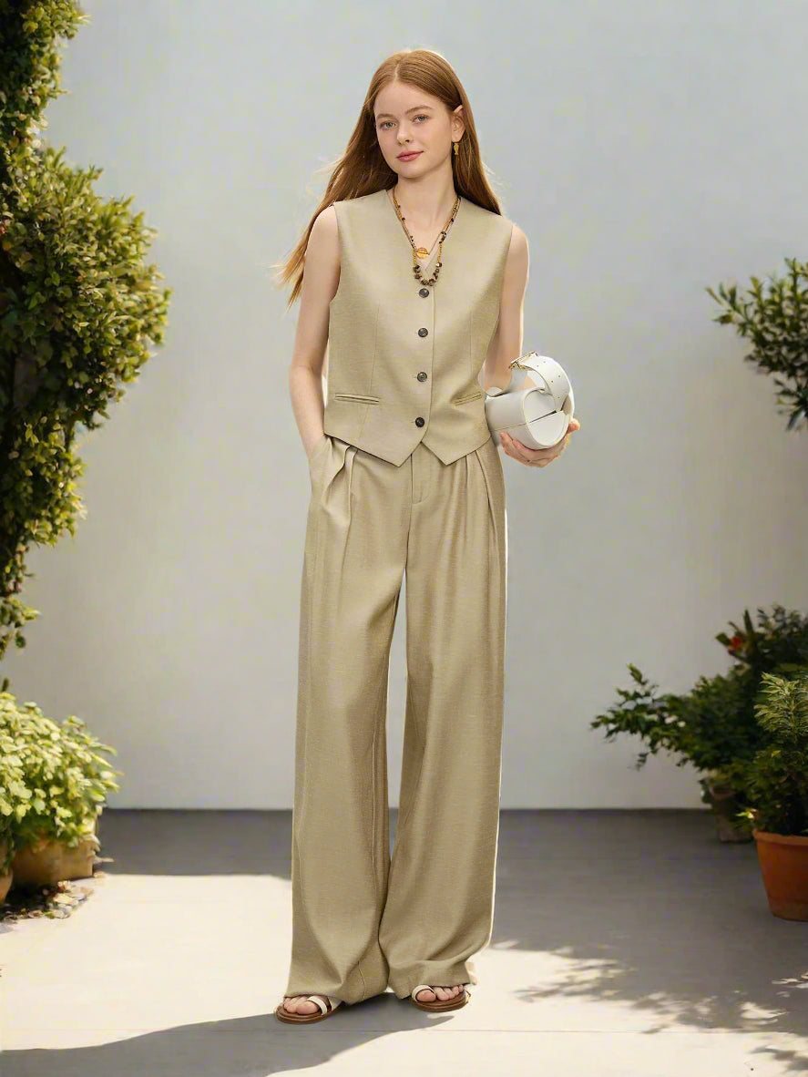 Woman wearing a beige vest and wide-leg pants holding a white handbag against a plain background