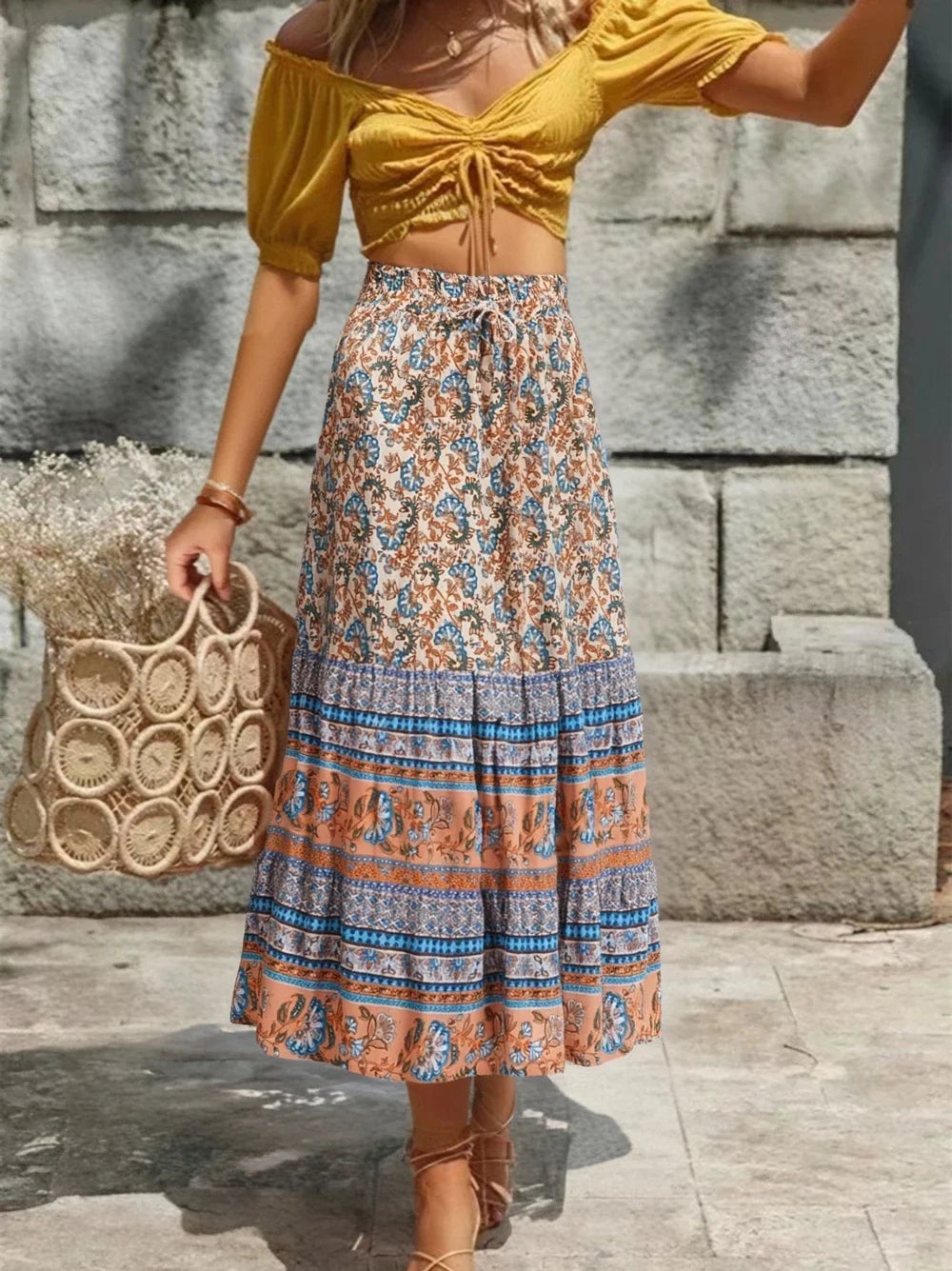 Woman wearing a yellow top and patterned skirt holding a woven bag, standing against a stone wall.