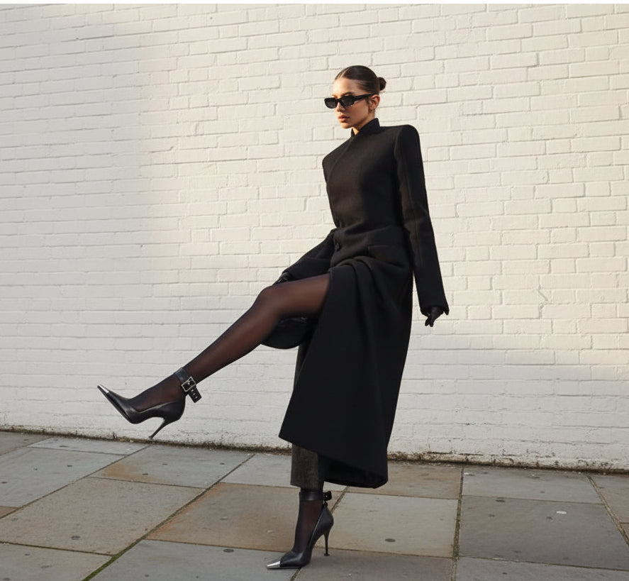Woman in a black outfit leaning against a white wall.