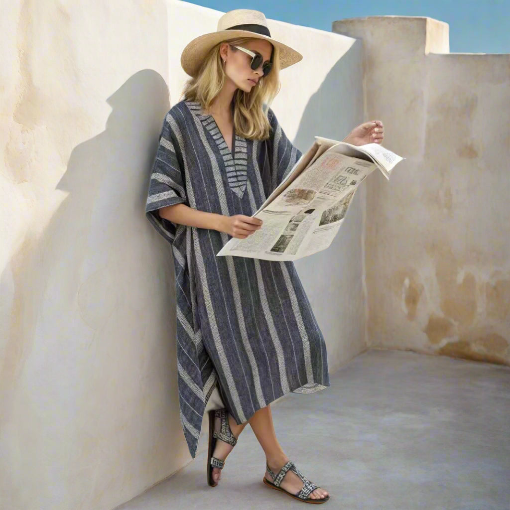 Woman in a striped dress and hat reading a newspaper against a white wall.