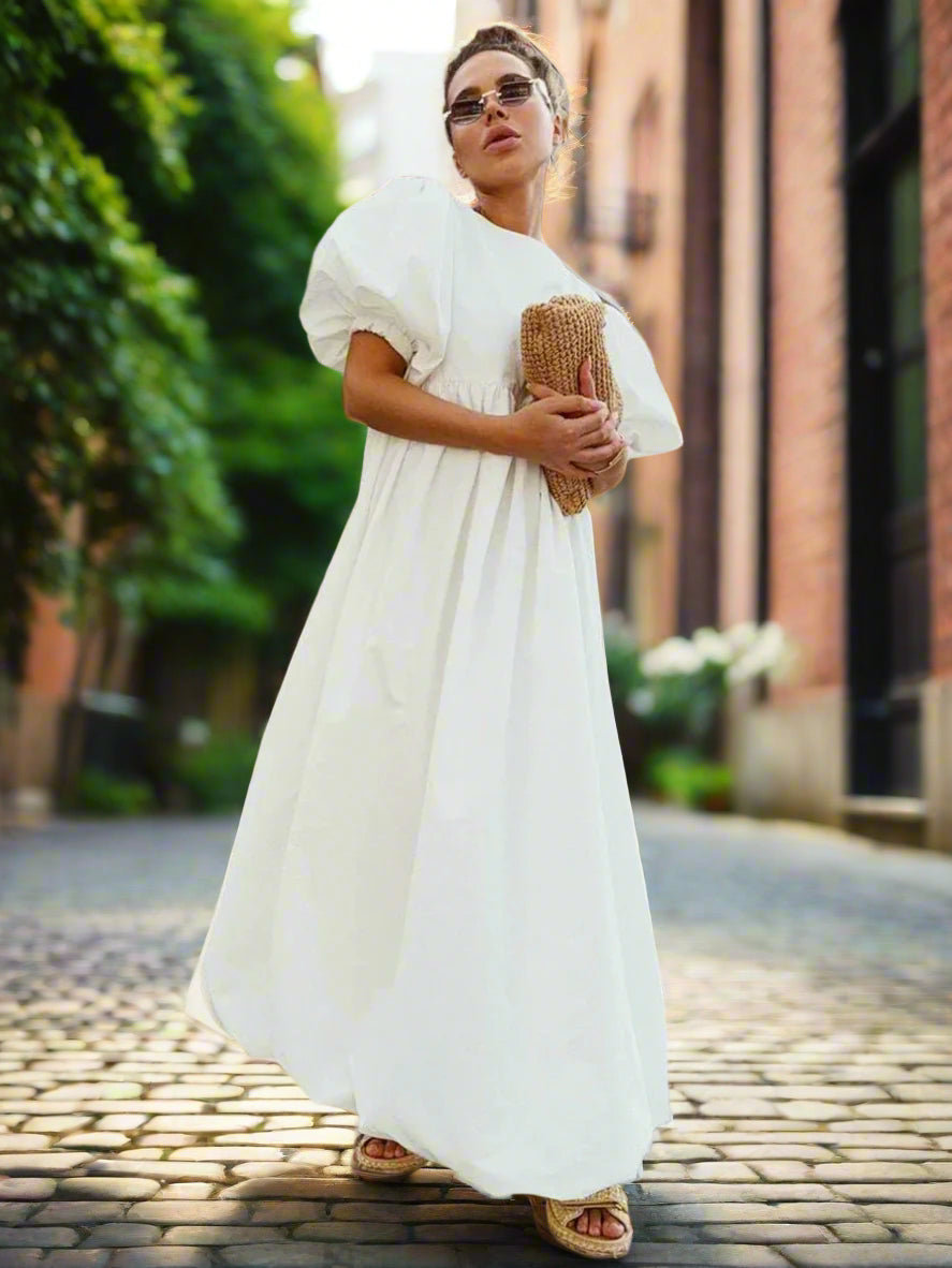 Woman in a white dress holding a woven bag against a backyard garden, sunny