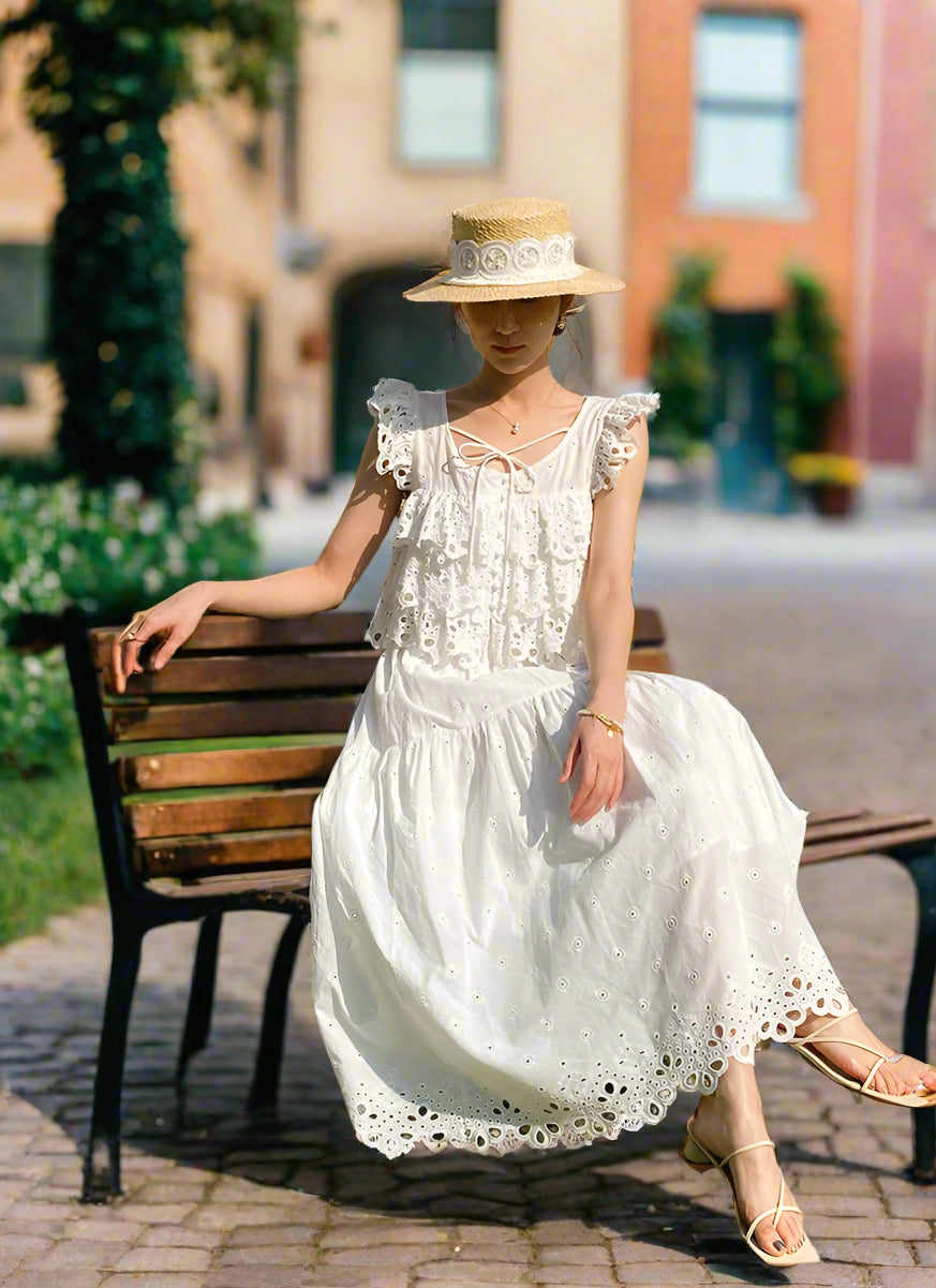 Woman in a white dress and straw hat sitting on a green and beige bench outdoors.
