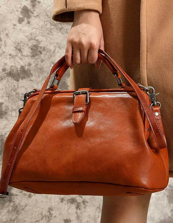 Brown leather handbag held by a person wearing a tan coat against a neutral background