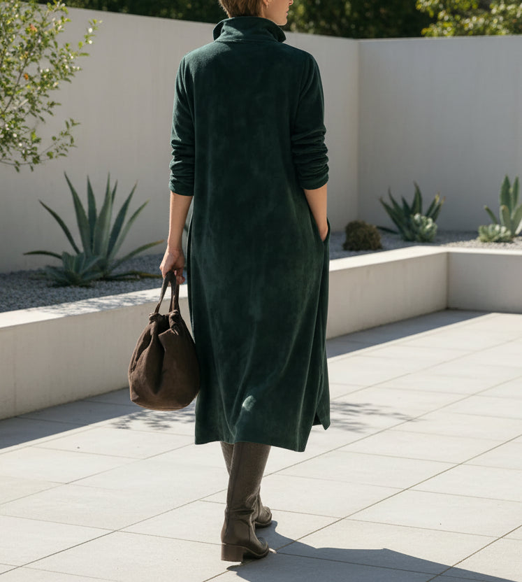 Person wearing a long green dress holding a brown bag against a minimalist backyard