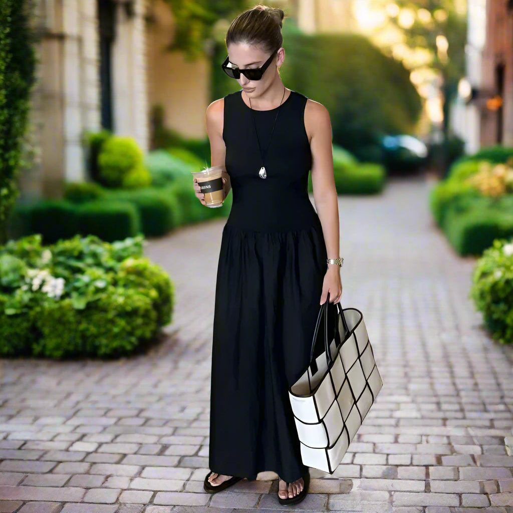 Woman in a black dress holding a white tote bag and a drink, standing in a backyard garden