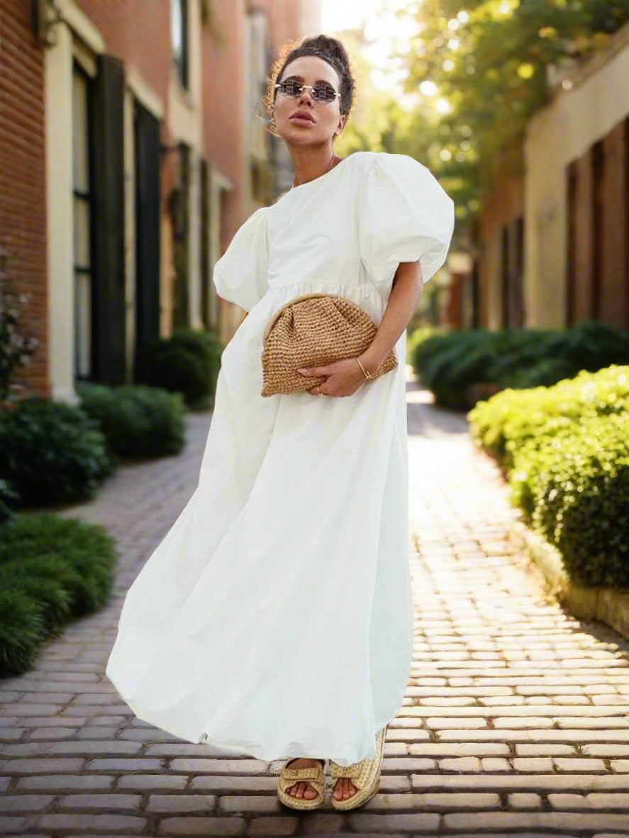 Woman in a white dress holding a woven handbag against a backyard garden, sunny