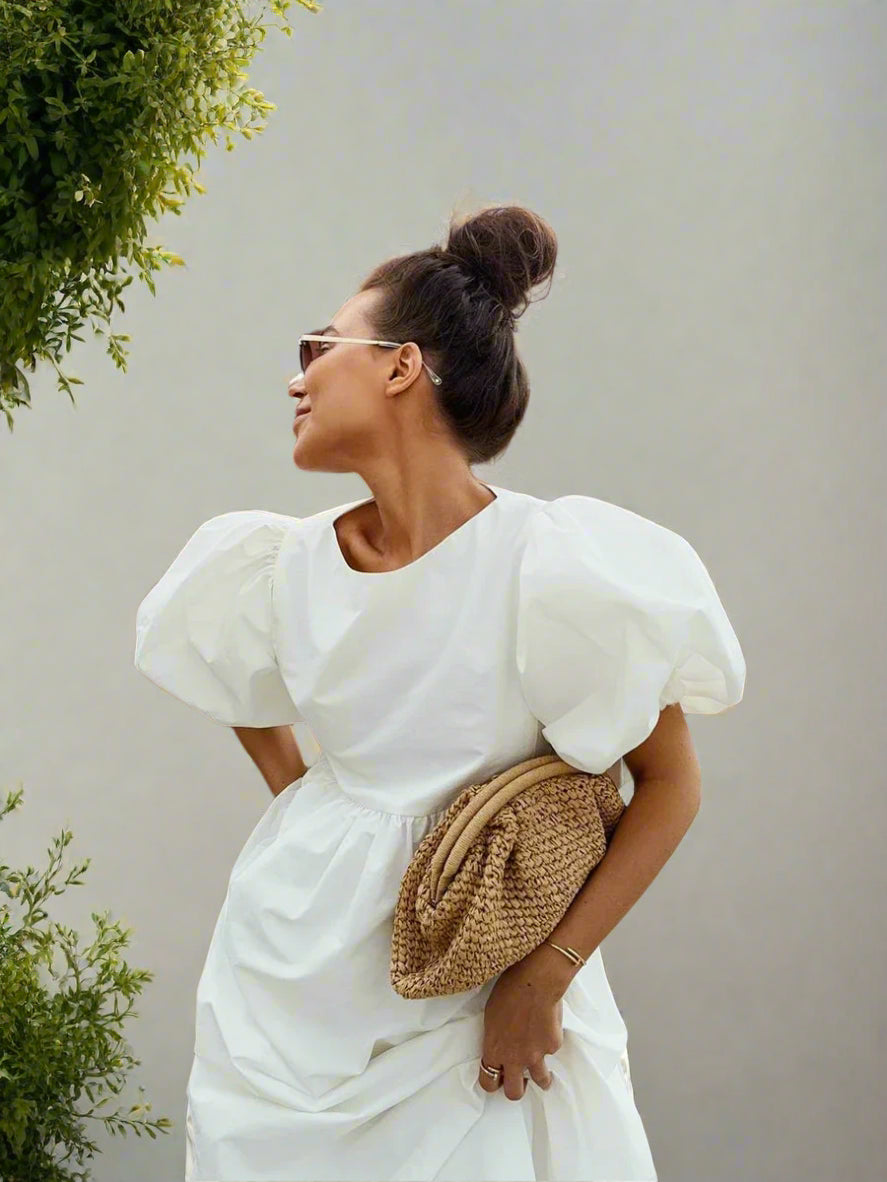 Woman in a white dress with puffed sleeves holding a straw clutch against a backyard garden, sunny