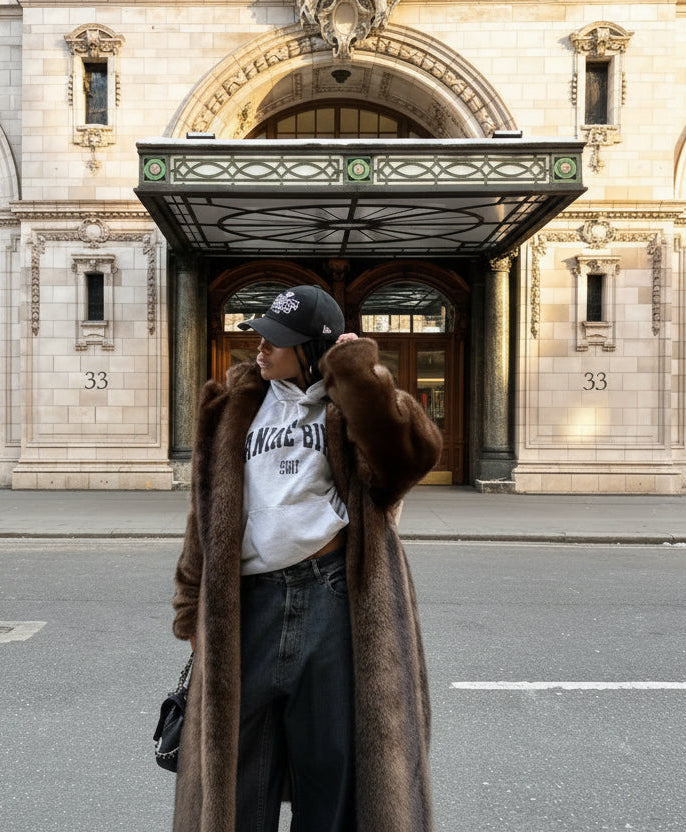 Person wearing a brown coat and white sweatshirt with text, standing in front of a large building.