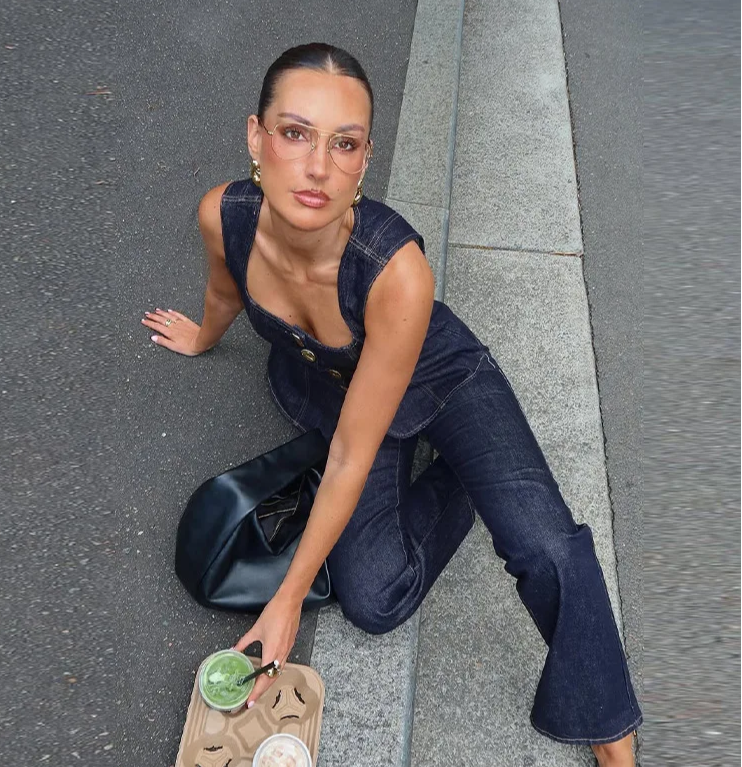 Woman in denim overalls sitting on a street with a drink in a paper cup.