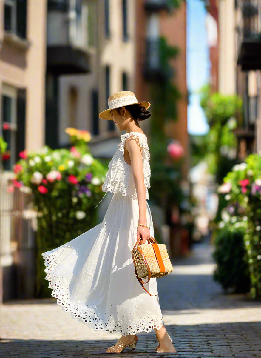 Woman in a white dress standing in front of a white wall with red flowers and a backyard garden, sunny